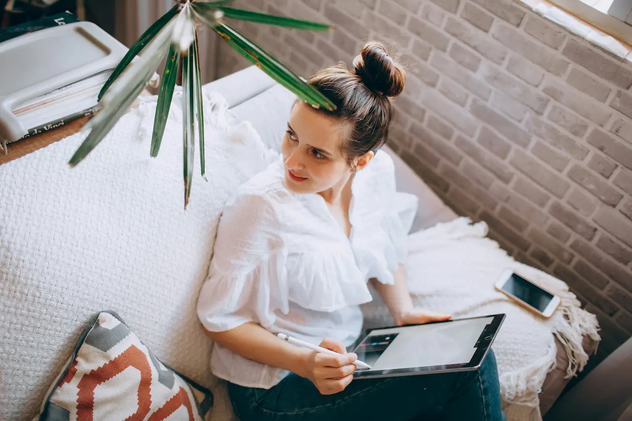 Woman relaxing on a couch using a tablet beside a bright window in a modern cozy interior.